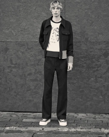 Full-length studio shot of a fair-skinned male adult model wearing dark jacket, cream sweater, dark pants, and sneakers against a grey wall.