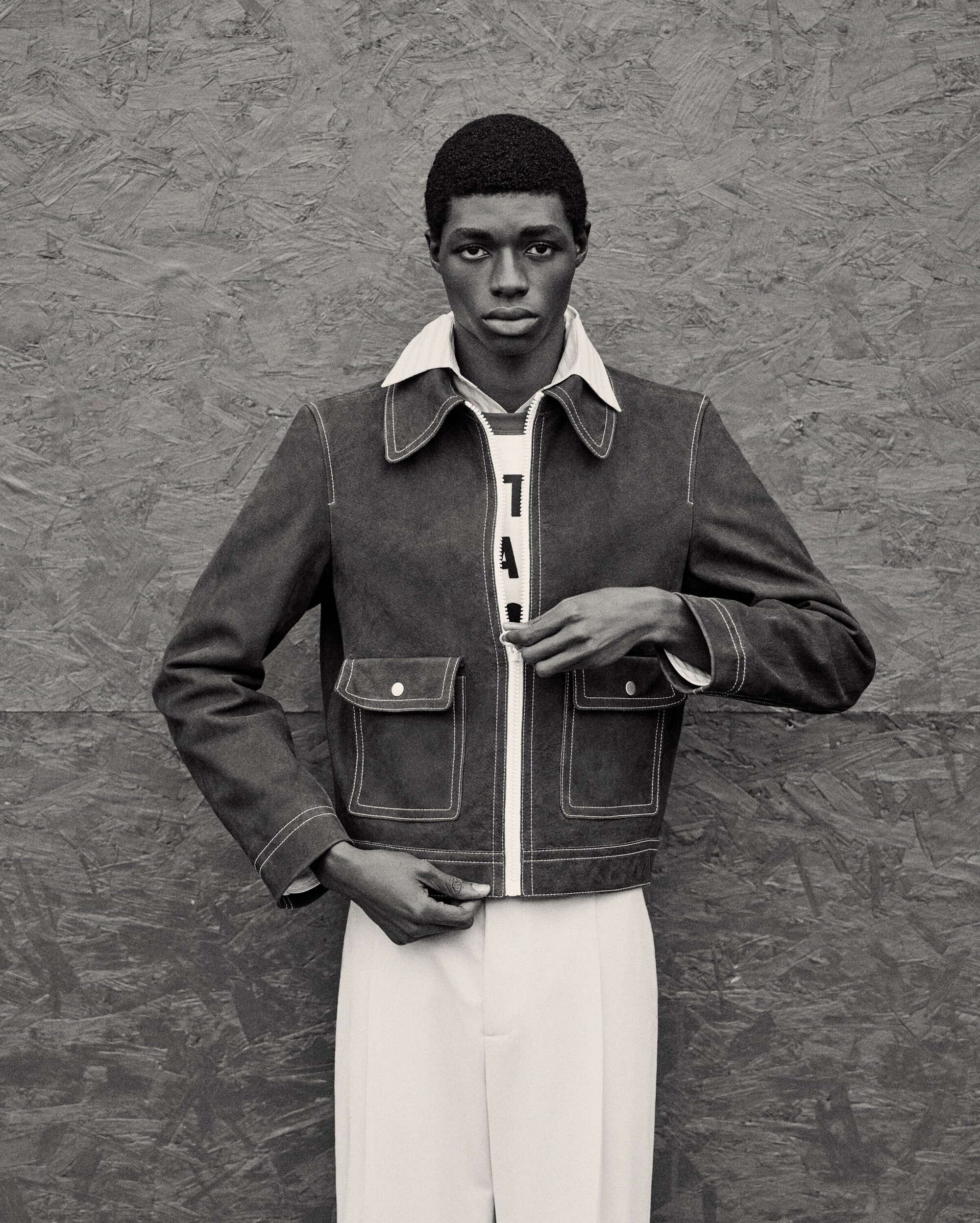 Portrait of a Black male model in a denim jacket with white trim, white pants, posing against a grey textured background.