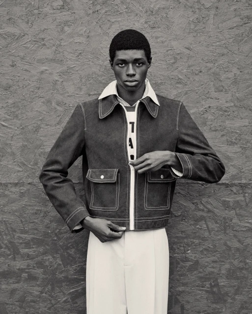 Portrait of a Black male model in a denim jacket with white trim, white pants, posing against a grey textured background.