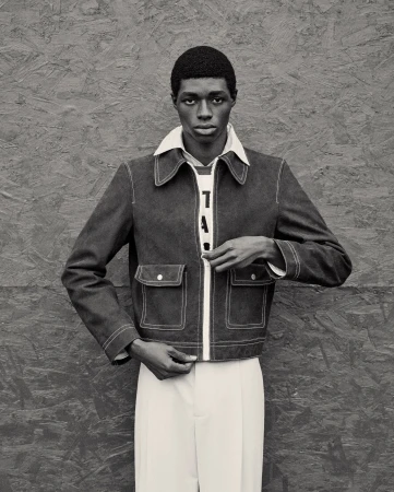 Portrait of a Black male model in a denim jacket with white trim, white pants, posing against a grey textured background.