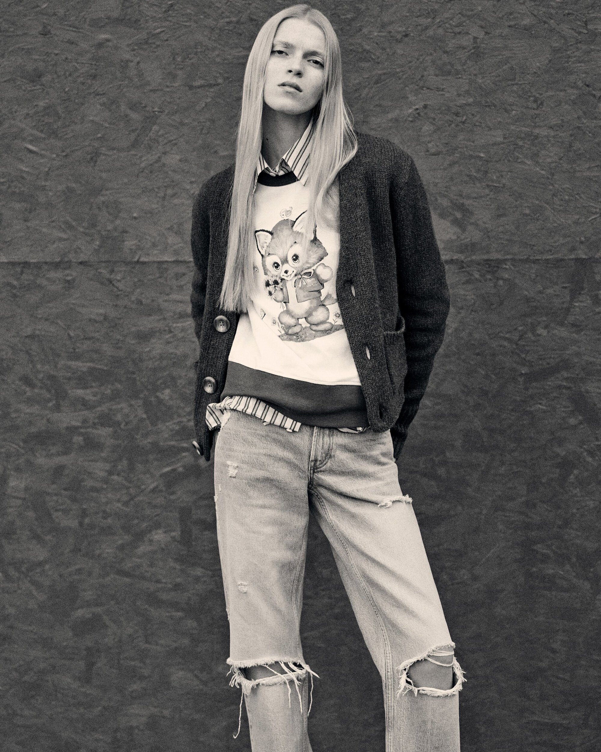 Blonde woman in a cartoon sweatshirt, cardigan, and ripped jeans posing against a grey textured background in a fashion editorial shoot.