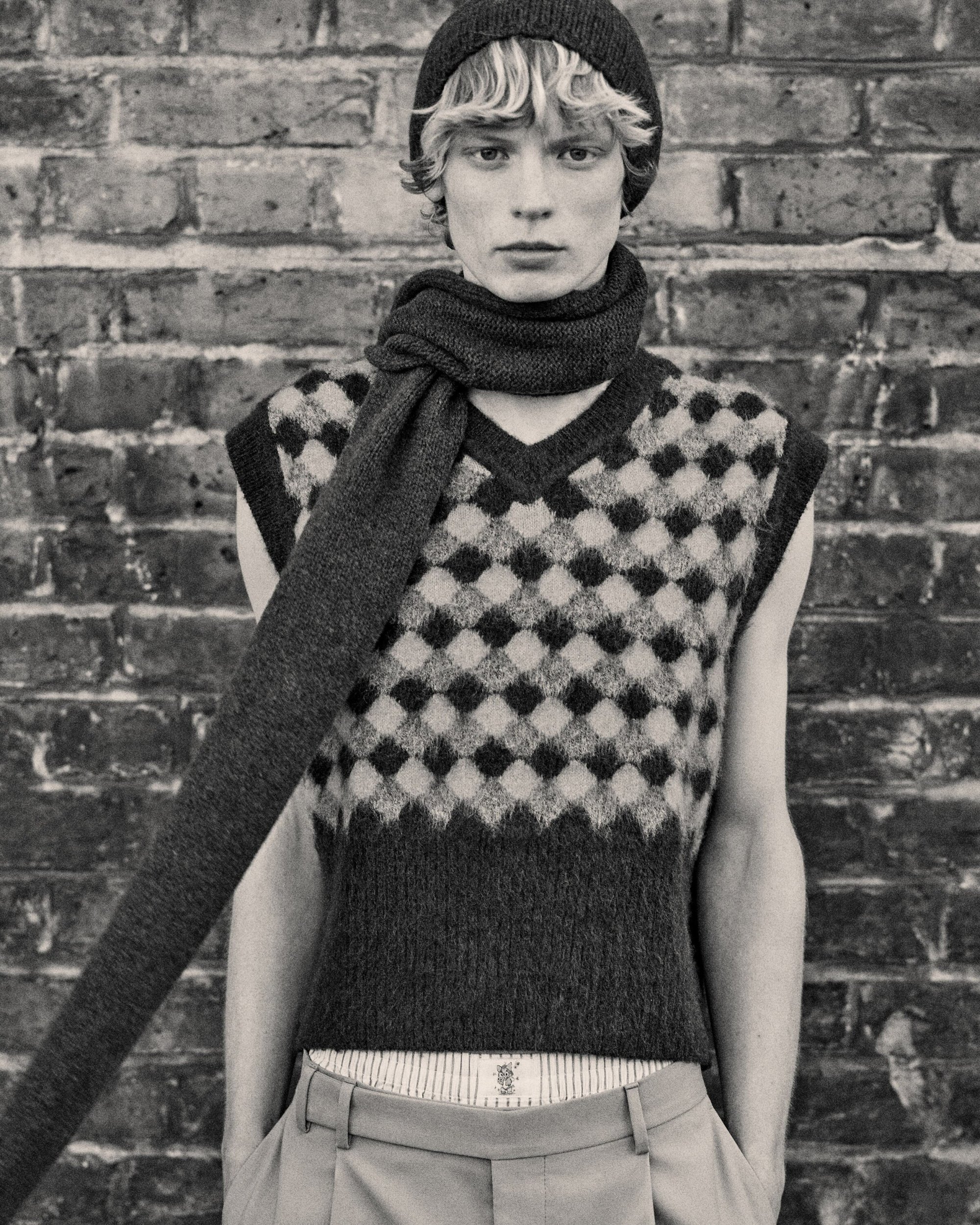 Monochrome portrait of a young male model in a vest, scarf, and beanie, posing against a brick wall in a fashion editorial style.