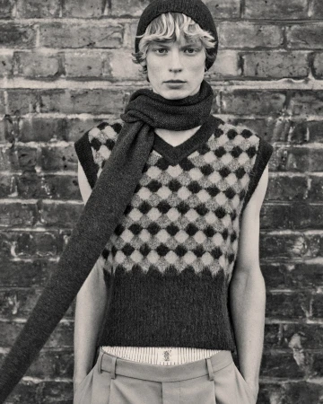 Monochrome portrait of a young male model in a vest, scarf, and beanie, posing against a brick wall in a fashion editorial style.