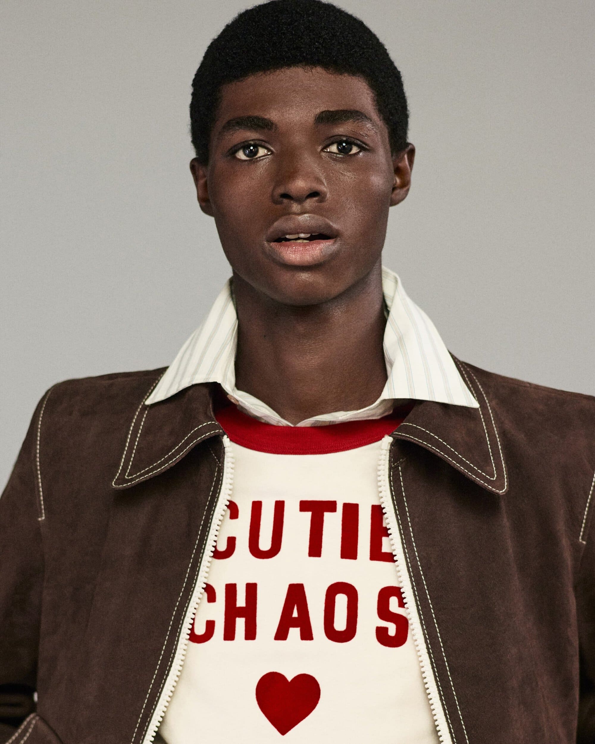 Close-up studio shot of a young adult Black male wearing a brown jacket and white shirt with red text graphic and heart design.