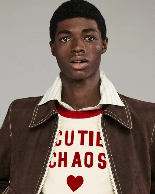 Close-up studio shot of a young adult Black male wearing a brown jacket and white shirt with red text graphic and heart design.