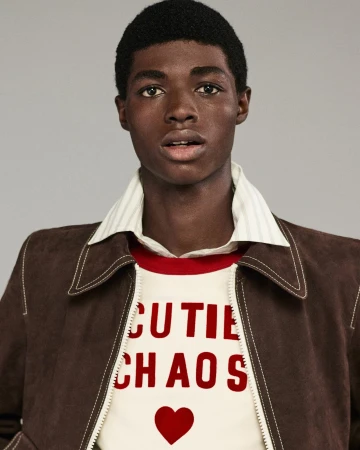 Close-up studio shot of a young adult Black male wearing a brown jacket and white shirt with red text graphic and heart design.