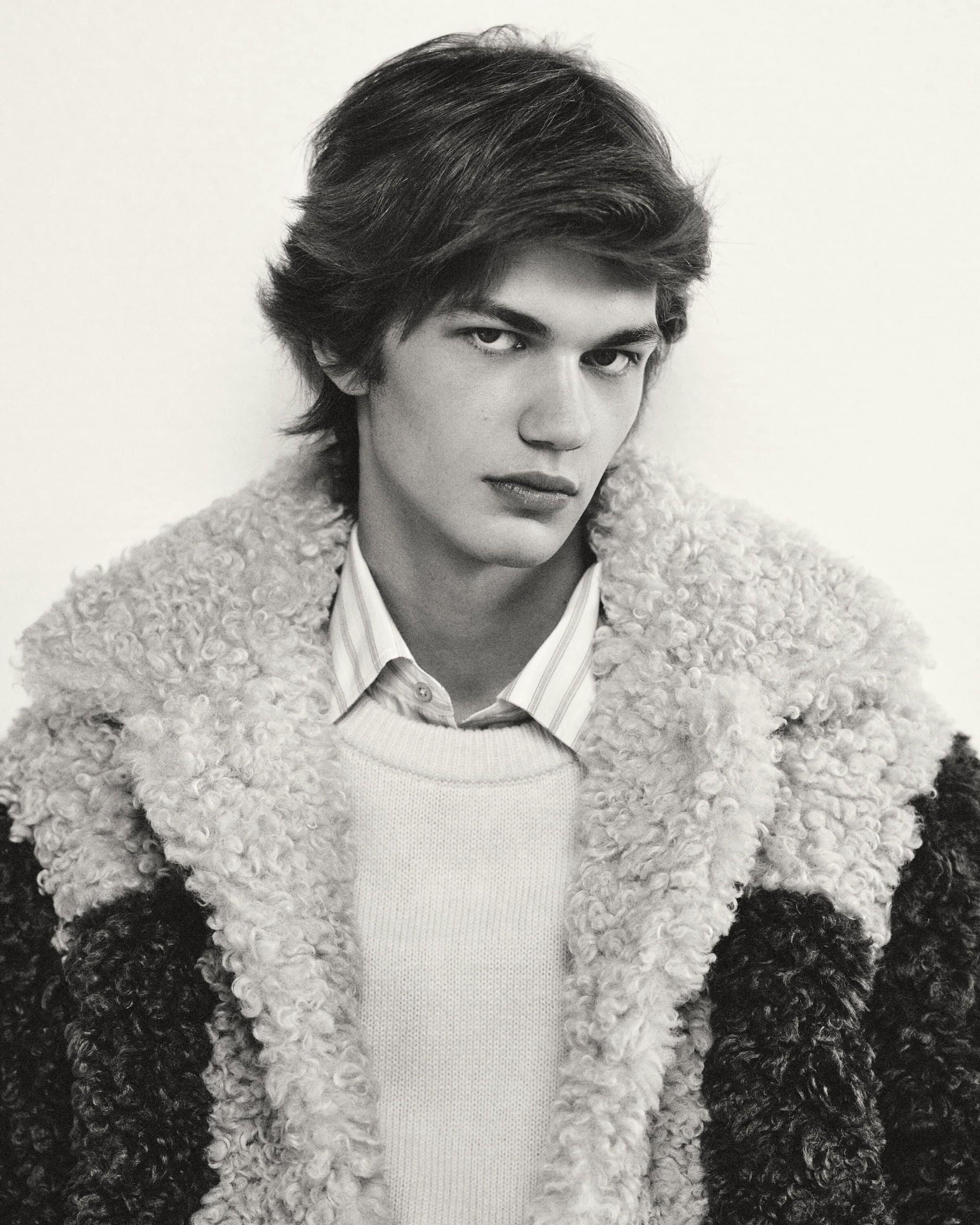 Monochromatic portrait of a fair-skinned young adult male wearing a sheepskin coat, with a neutral expression against a plain background.