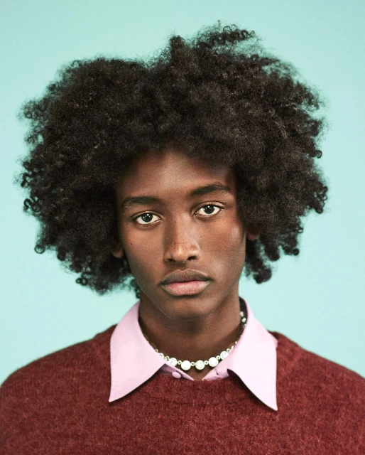 Portrait of a young Black male model with an Afro, wearing a maroon sweater and pearl necklace, against a light blue background.