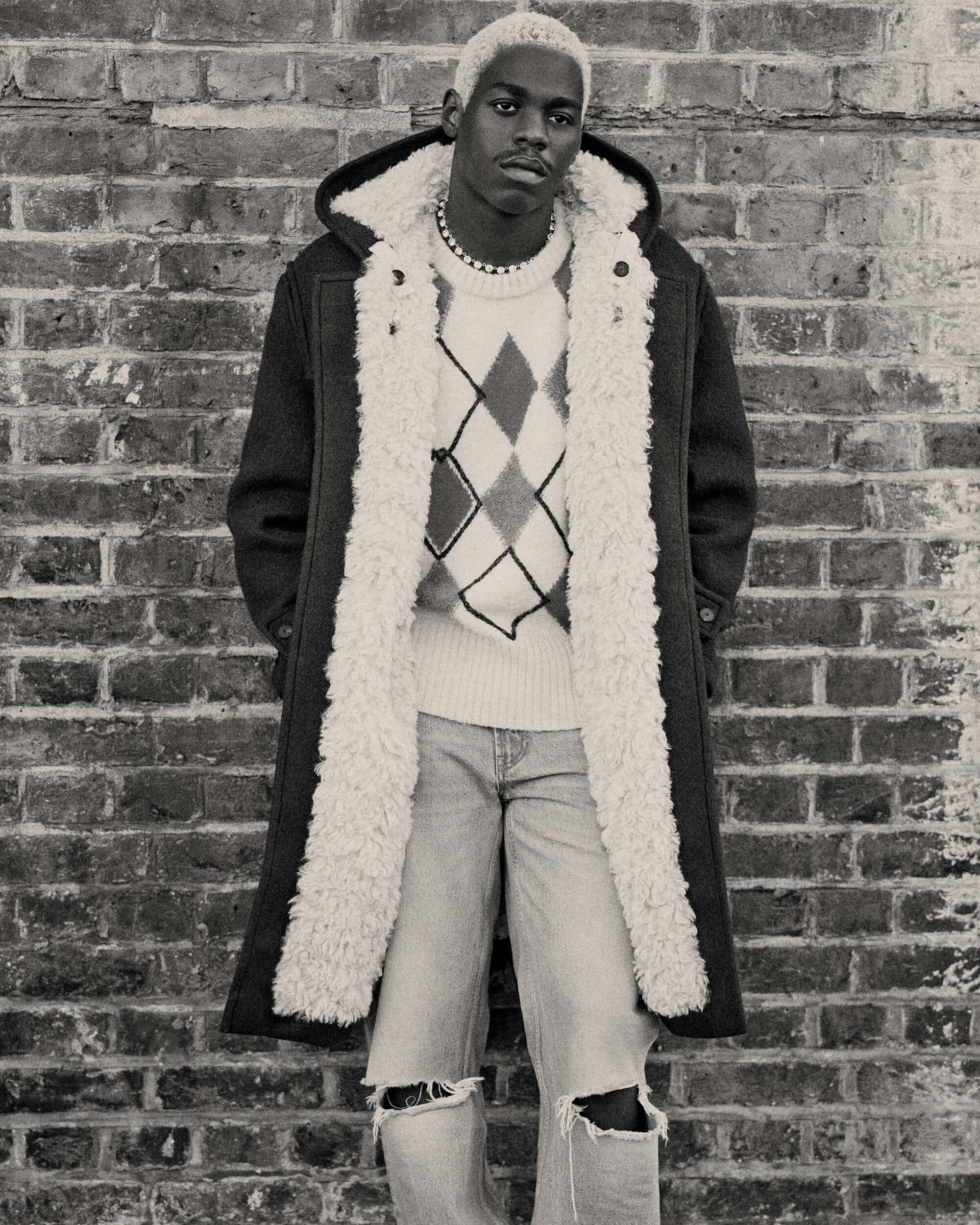 Monochrome portrait of a black male with bleached hair in an argyle sweater, distressed jeans, and a fur-lined coat standing against a brick wall.