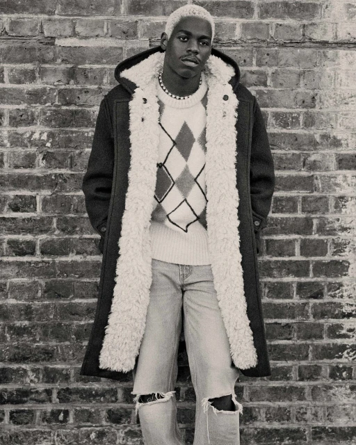 Monochrome portrait of a black male with bleached hair in an argyle sweater, distressed jeans, and a fur-lined coat standing against a brick wall.