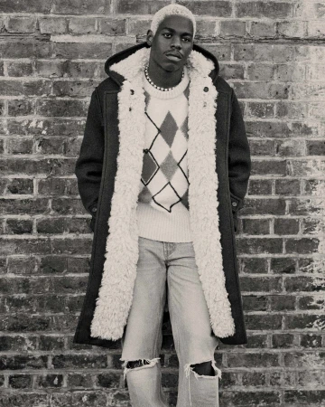 Monochrome portrait of a black male with bleached hair in an argyle sweater, distressed jeans, and a fur-lined coat standing against a brick wall.