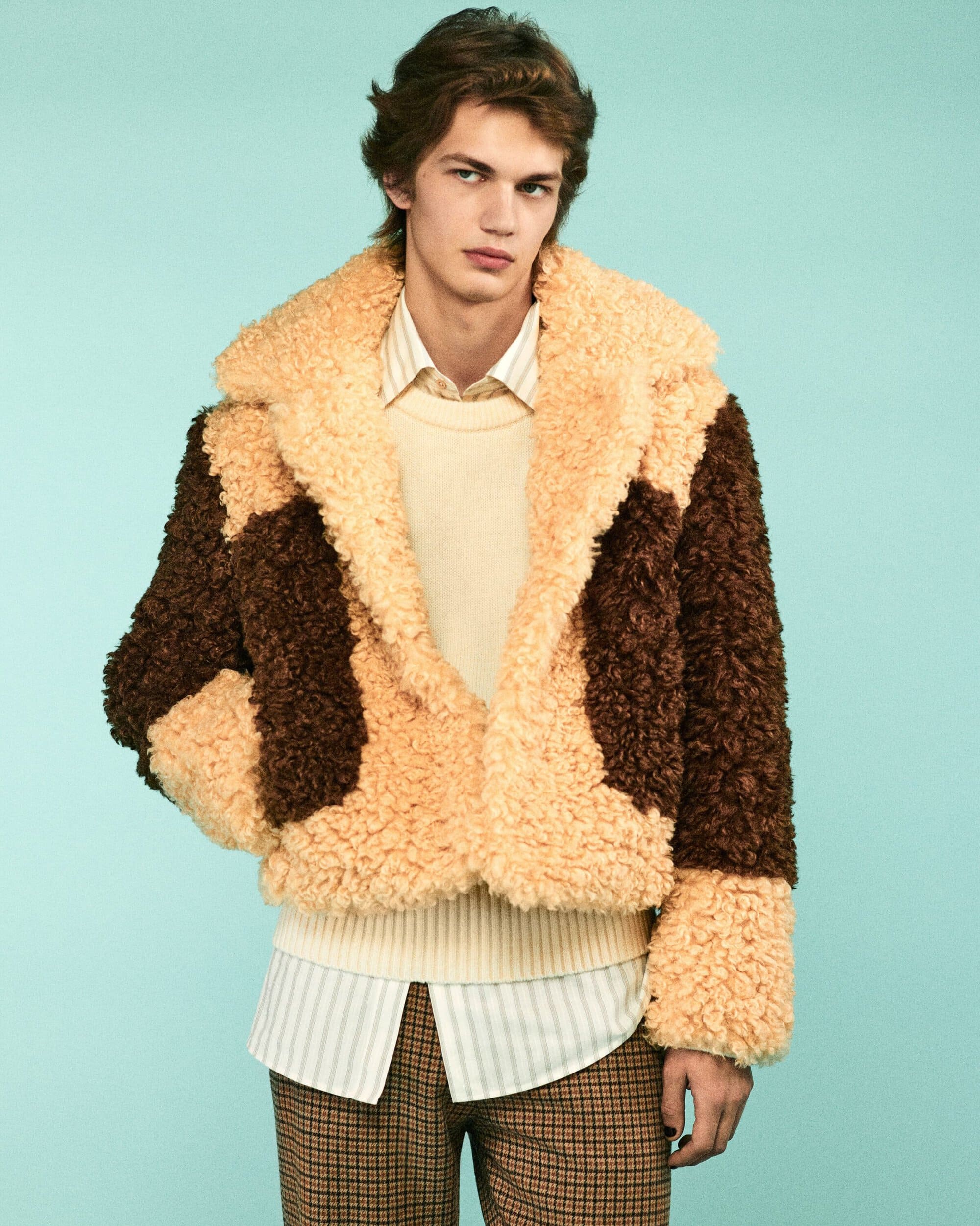 Young adult man posing indoors wearing a brown and tan fleece jacket, striped shirt, and plaid pants against a blue background.