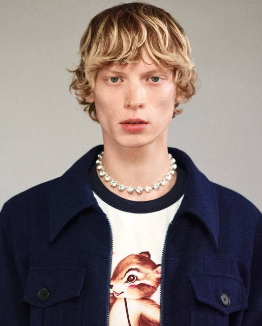 Man with blond wavy hair wearing a navy jacket, white t-shirt with a cartoon squirrel, and a diamond necklace indoors.