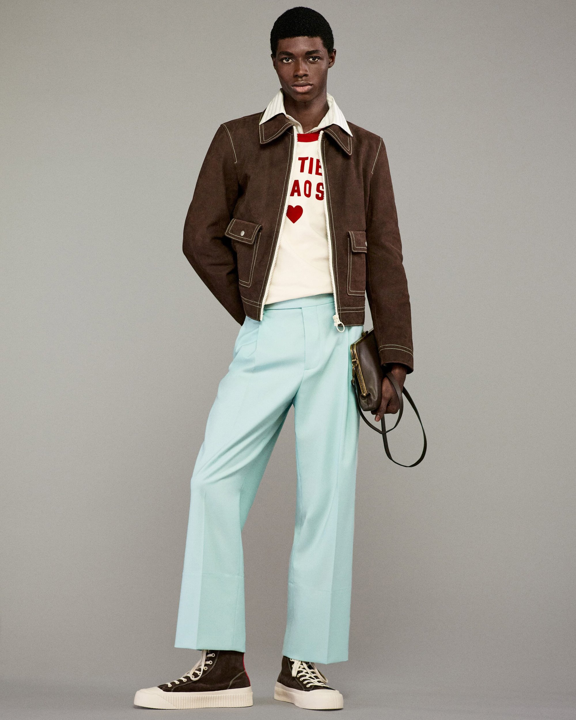 A single Black male model stands in a studio wearing a brown jacket, white top, and blue pants, holding a dark brown bag, and wearing high-tops.