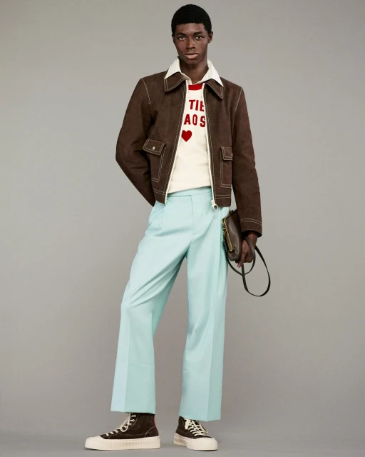 A single Black male model stands in a studio wearing a brown jacket, white top, and blue pants, holding a dark brown bag, and wearing high-tops.