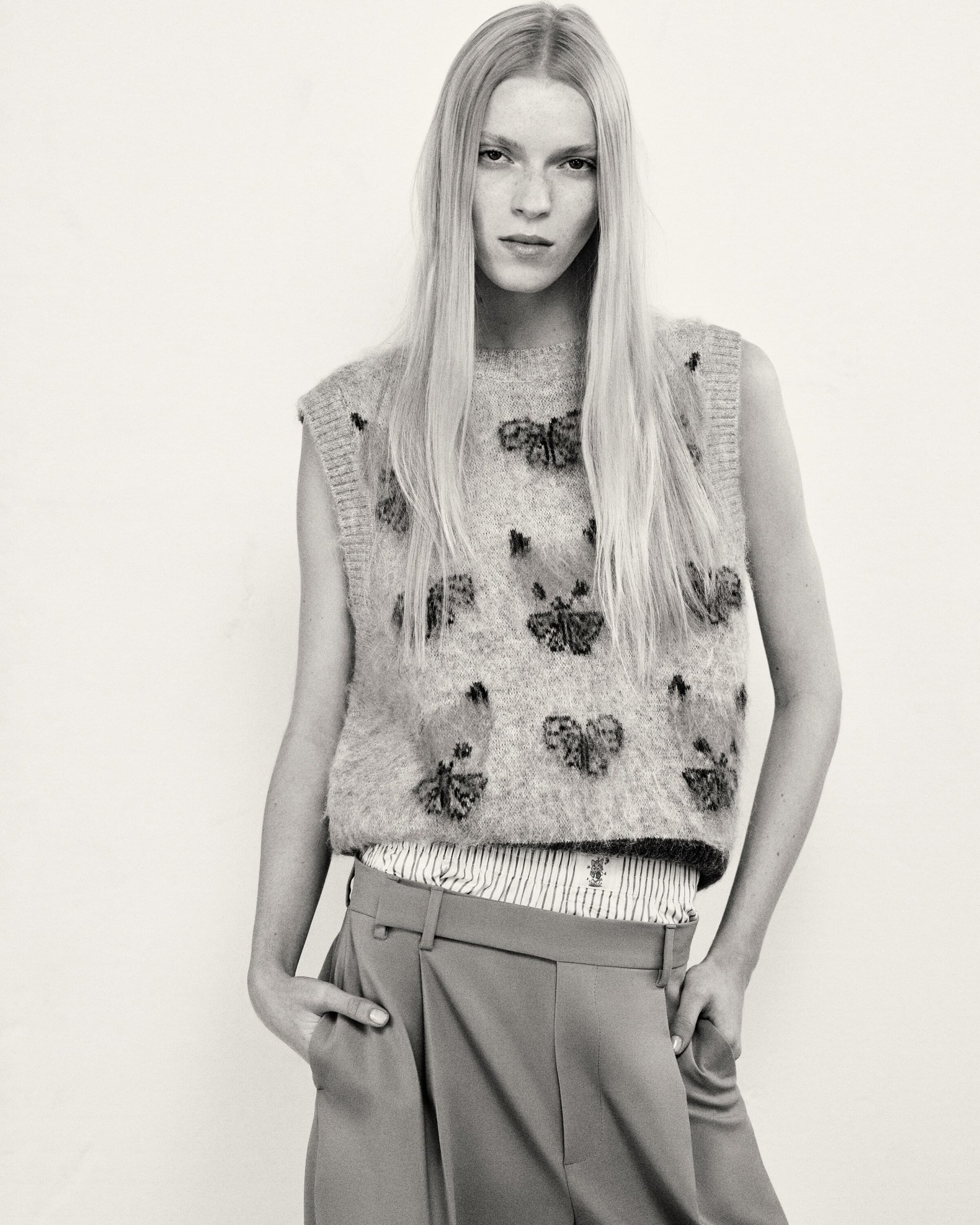 A fair-skinned woman with long blonde hair models a sleeveless butterfly-embroidered sweater and pleated trousers in a studio shot.
