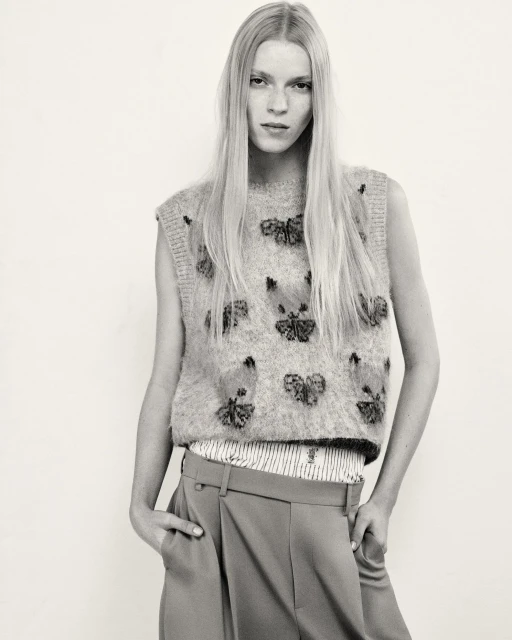A fair-skinned woman with long blonde hair models a sleeveless butterfly-embroidered sweater and pleated trousers in a studio shot.