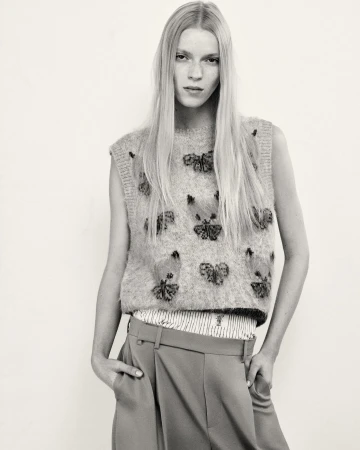 A fair-skinned woman with long blonde hair models a sleeveless butterfly-embroidered sweater and pleated trousers in a studio shot.