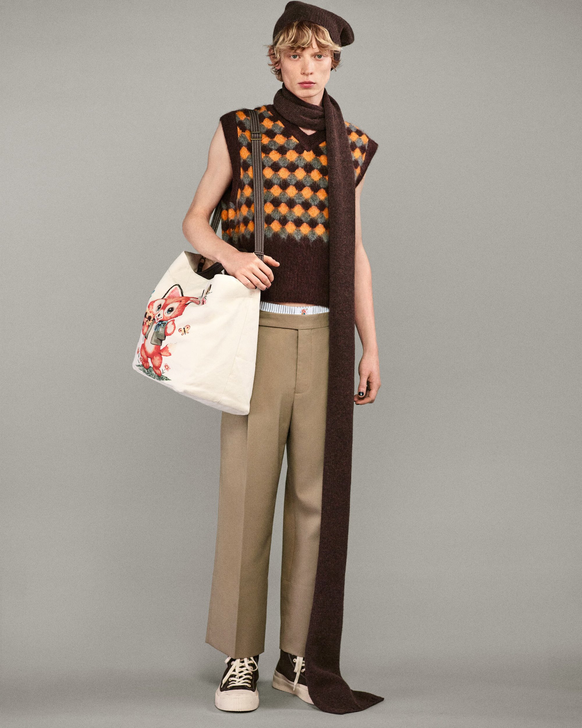 Young adult in patterned vest, brown scarf, and beret carrying illustrated tote bag in a full studio shot on grey background.