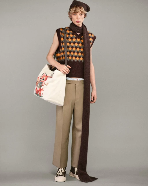 Young adult in patterned vest, brown scarf, and beret carrying illustrated tote bag in a full studio shot on grey background.
