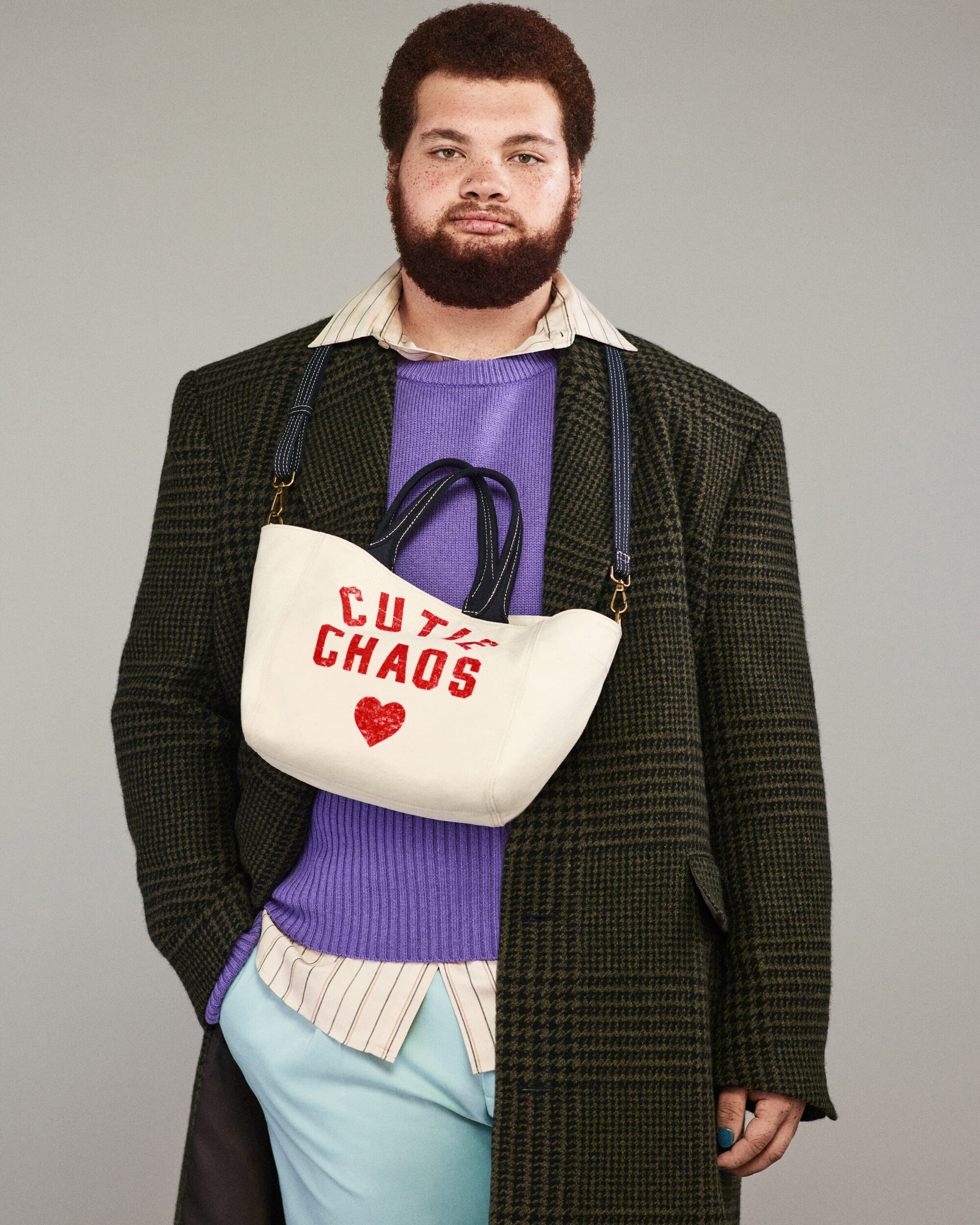 Man wearing a green plaid overcoat, purple sweater, and light blue pants, holding a white purse that reads 'Cutie Chaos'.
