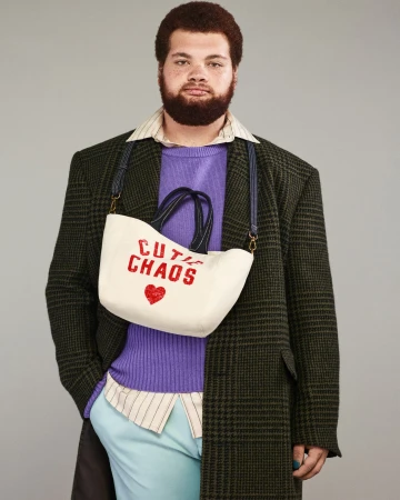 Man wearing a green plaid overcoat, purple sweater, and light blue pants, holding a white purse that reads 'Cutie Chaos'.