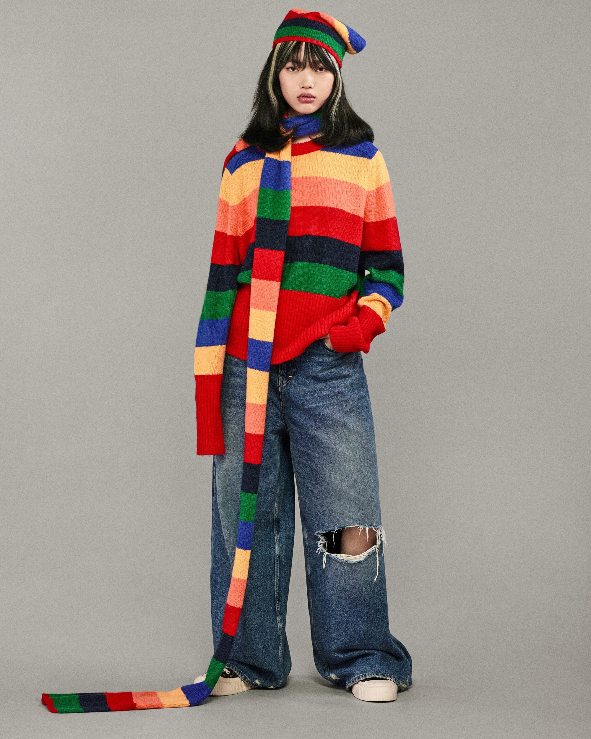 A young woman poses in a colorful striped sweater, scarf, and hat ensemble with ripped jeans against a neutral studio backdrop.