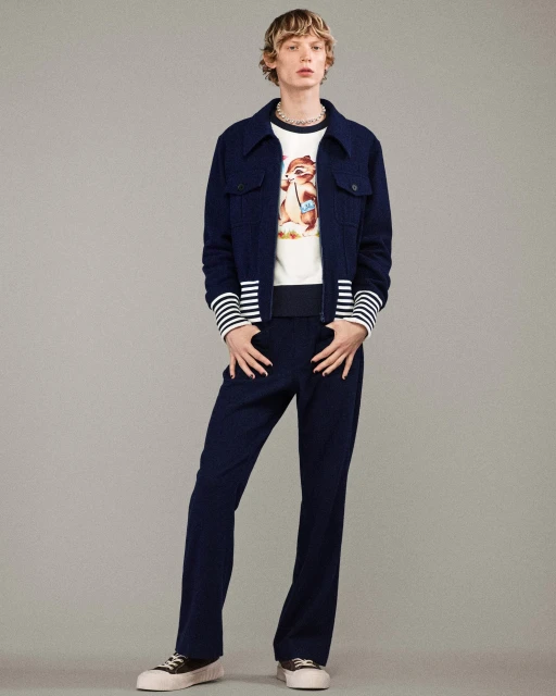 Full-body studio portrait of a young adult with fair skin and short, blonde hair wearing a matching navy jacket and pants ensemble.