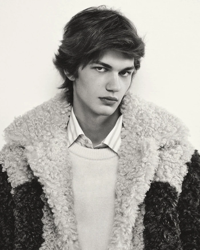 Portrait of a young man wearing a textured shearling collar coat and striped shirt against a plain white background.