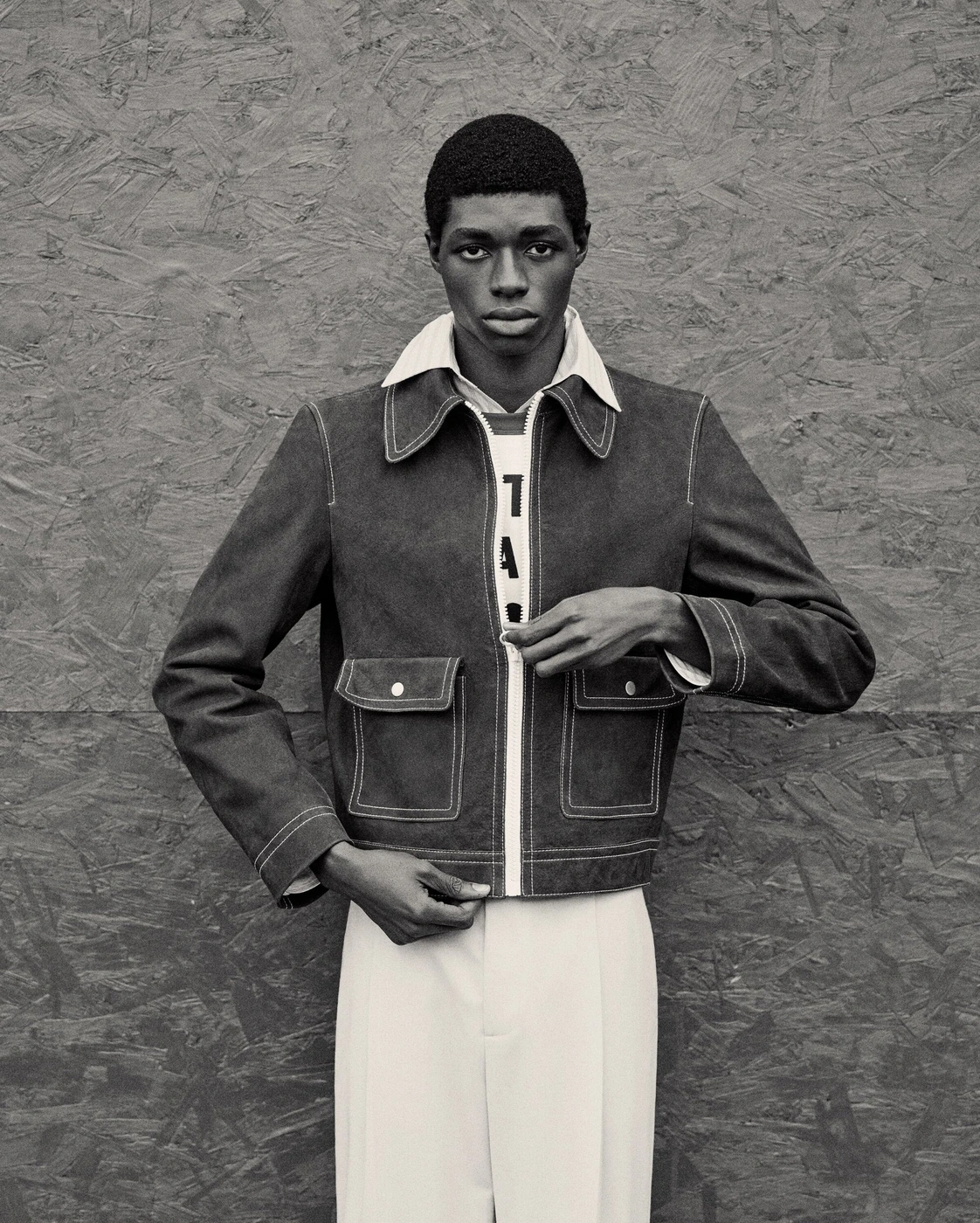 Black man wearing a zipped jacket and collared shirt posing against a textured wooden background