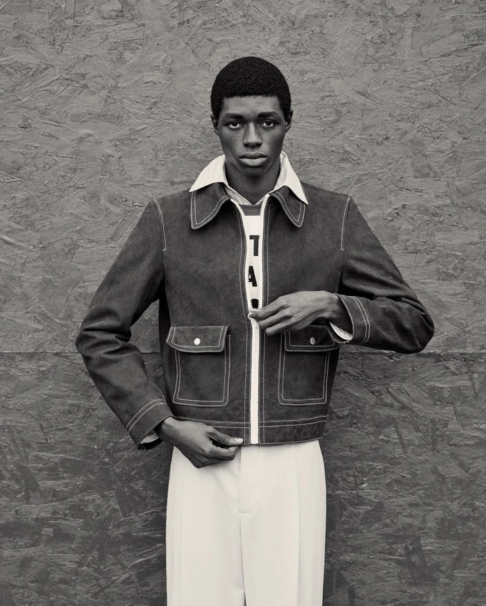 Black man wearing a zipped jacket and collared shirt posing against a textured wooden background