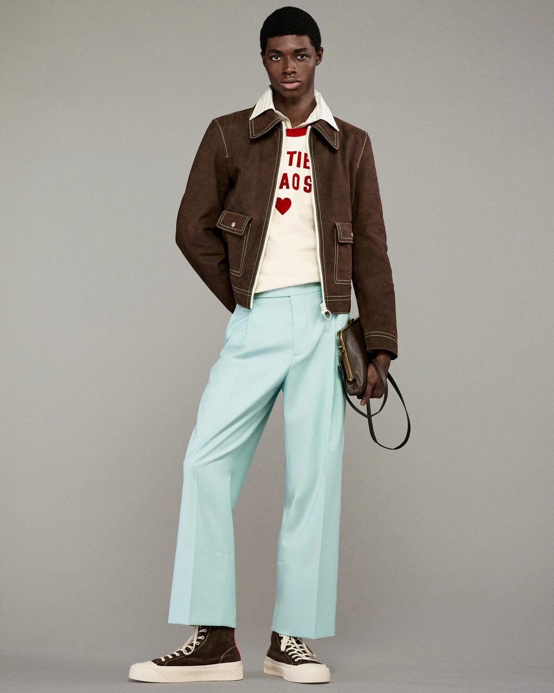 A male model wearing a brown jacket, light blue trousers, and dark sneakers against a grey seamless studio backdrop.