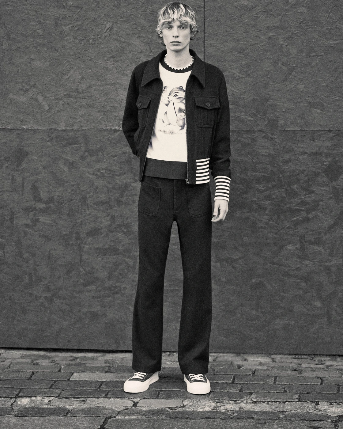 Monochrome fashion portrait of a male model wearing a dark jacket and trousers against a textured background.