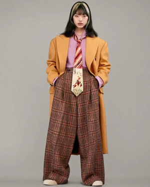 Model wearing an oversized mustard yellow coat, purple shirt, patterned tie, and plaid trousers against grey background