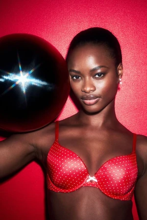 Portrait of an adult woman with dark skin, wearing a red polka-dot bra and holding a black ball against a matching red background.