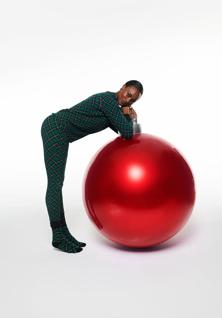 Woman in plaid pajamas poses with a large, reflective red Christmas ornament against a white backdrop in a studio setting.