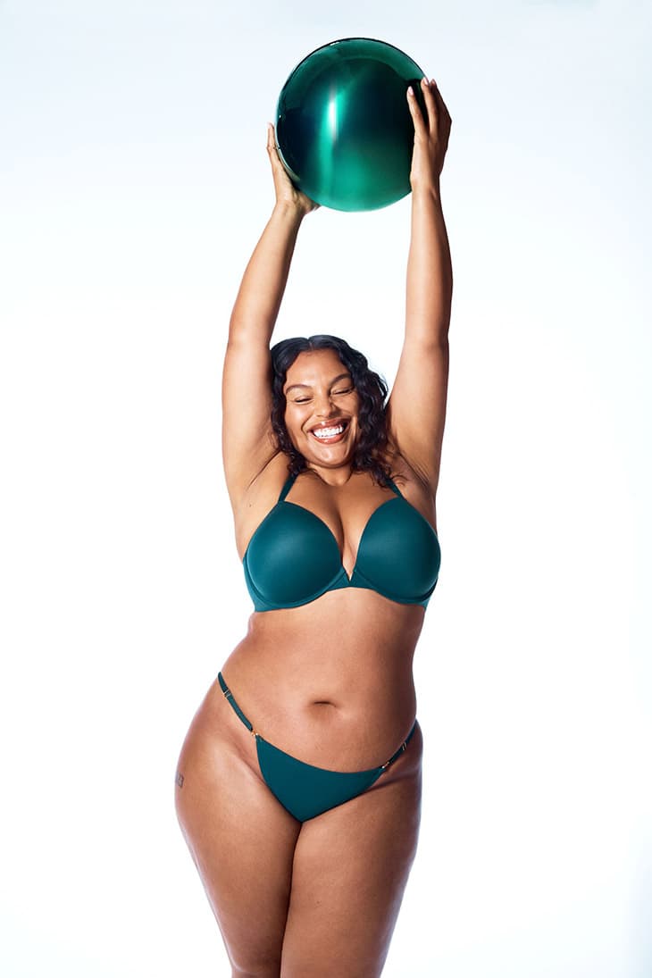 A curvy adult woman smiles while holding a metallic green ball above her head in a studio shot against a white backdrop.
