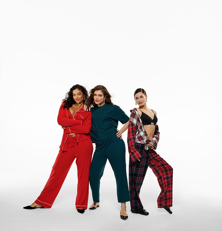Three women pose in a photo studio, wearing red, teal, and plaid pajama-style outfits, standing against a white backdrop for a fashion shoot.