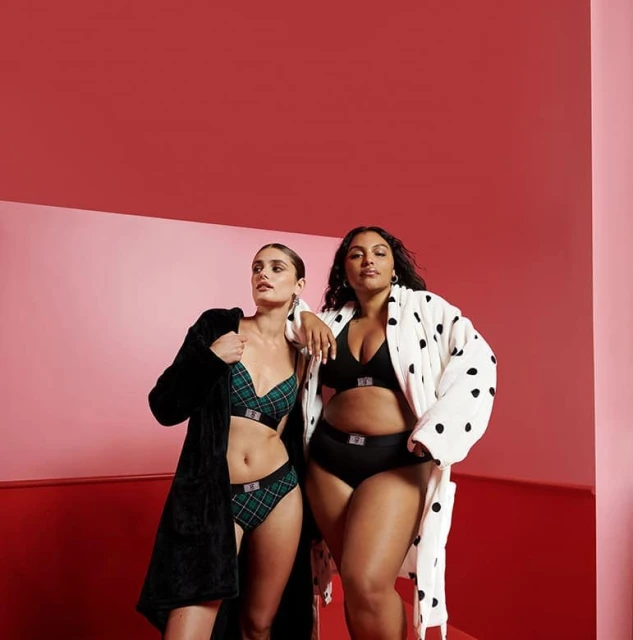 Two women pose in a photo studio wearing lingerie. One woman wears a green plaid set, while the other wears a black bra and panty set.