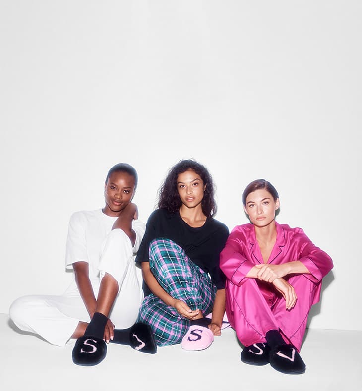 Three women sit in a studio wearing fashionable loungewear and slippers. The backdrop is a bright white, creating a clean, modern look.