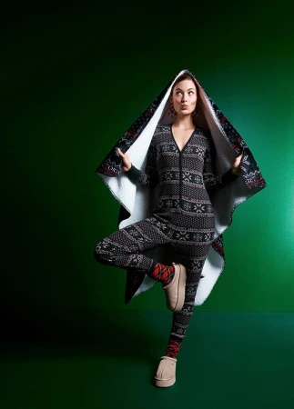 A woman in a patterned onesie and hooded cape poses in front of a green background, balancing on one leg while looking upwards.