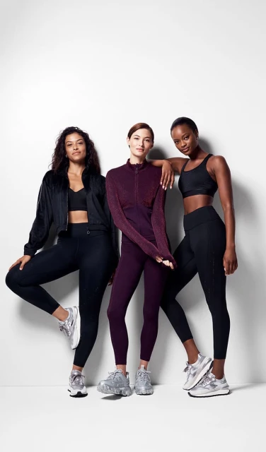 Three women in athletic wear pose against a white wall, showcasing fitness apparel. The subjects are wearing workout leggings and tops, and athletic shoes.