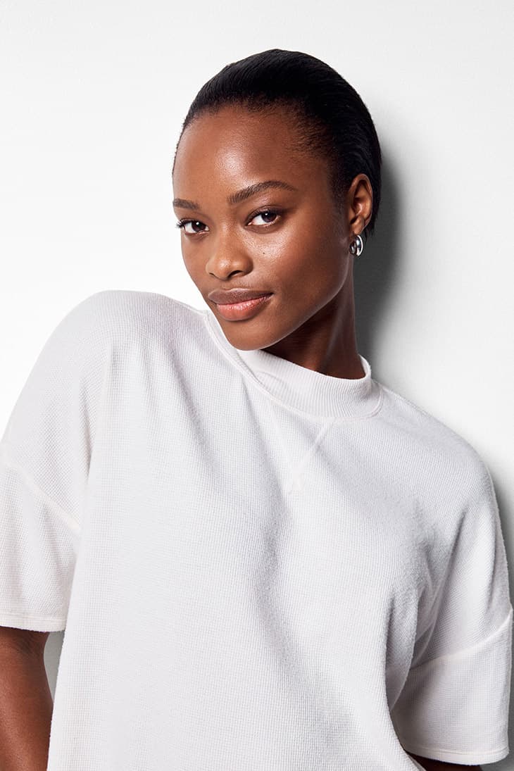 Close-up studio portrait of a Black woman with slicked-back hair, wearing a white textured t-shirt and silver hoop earrings, smiling slightly.