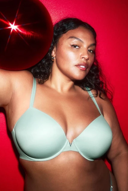 Close-up of a woman with curly dark hair and light skin wearing a light green bra against a red background with a red round object.