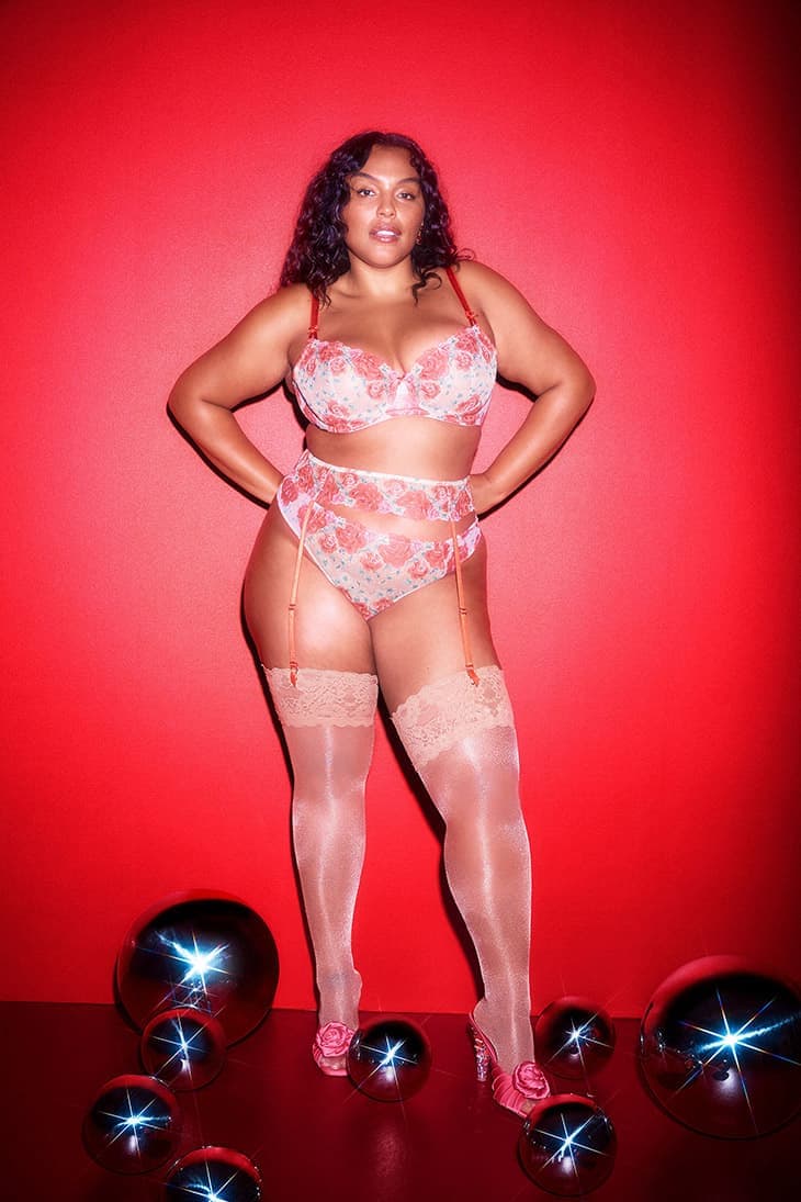 Full-length shot of a woman posing in lingerie on a red background, accented by metallic spheres, showcasing fashion and beauty.