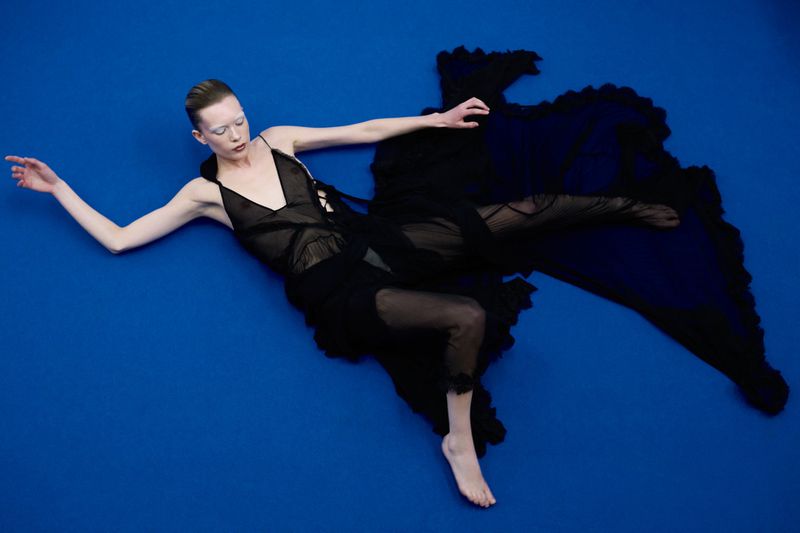 A slender adult female lies on a vibrant blue surface, adorned in a black sheer dress, striking a graceful pose with outstretched arms.