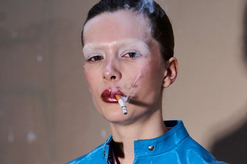 Close-up of a female model with striking white eyebrows and glossy red lips, smoking a cigarette in a neutral-toned indoor environment.