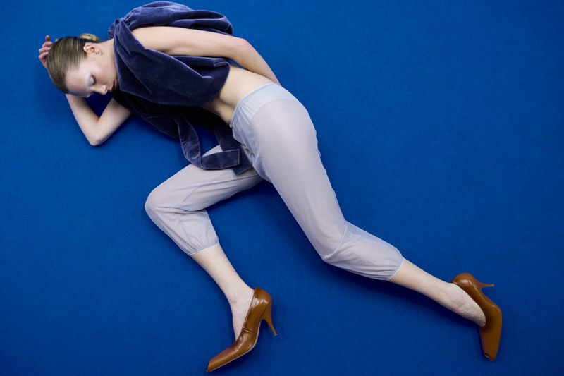 Model lying on blue background wearing velvet top and light blue pants with brown high heels in studio shot, top angle.