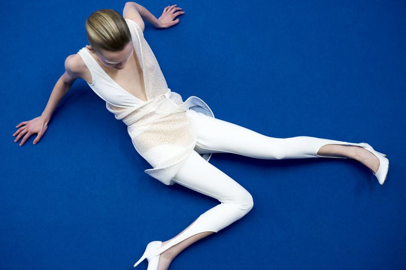Overhead shot of a fashion model in a white top, leggings, and heels lying on a vibrant blue surface in a dynamic pose.