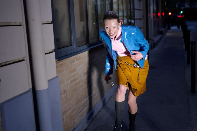 Smiling woman in blue jacket and gold skirt poses on a city street at night, highlighted by artificial light and visible car headlights.